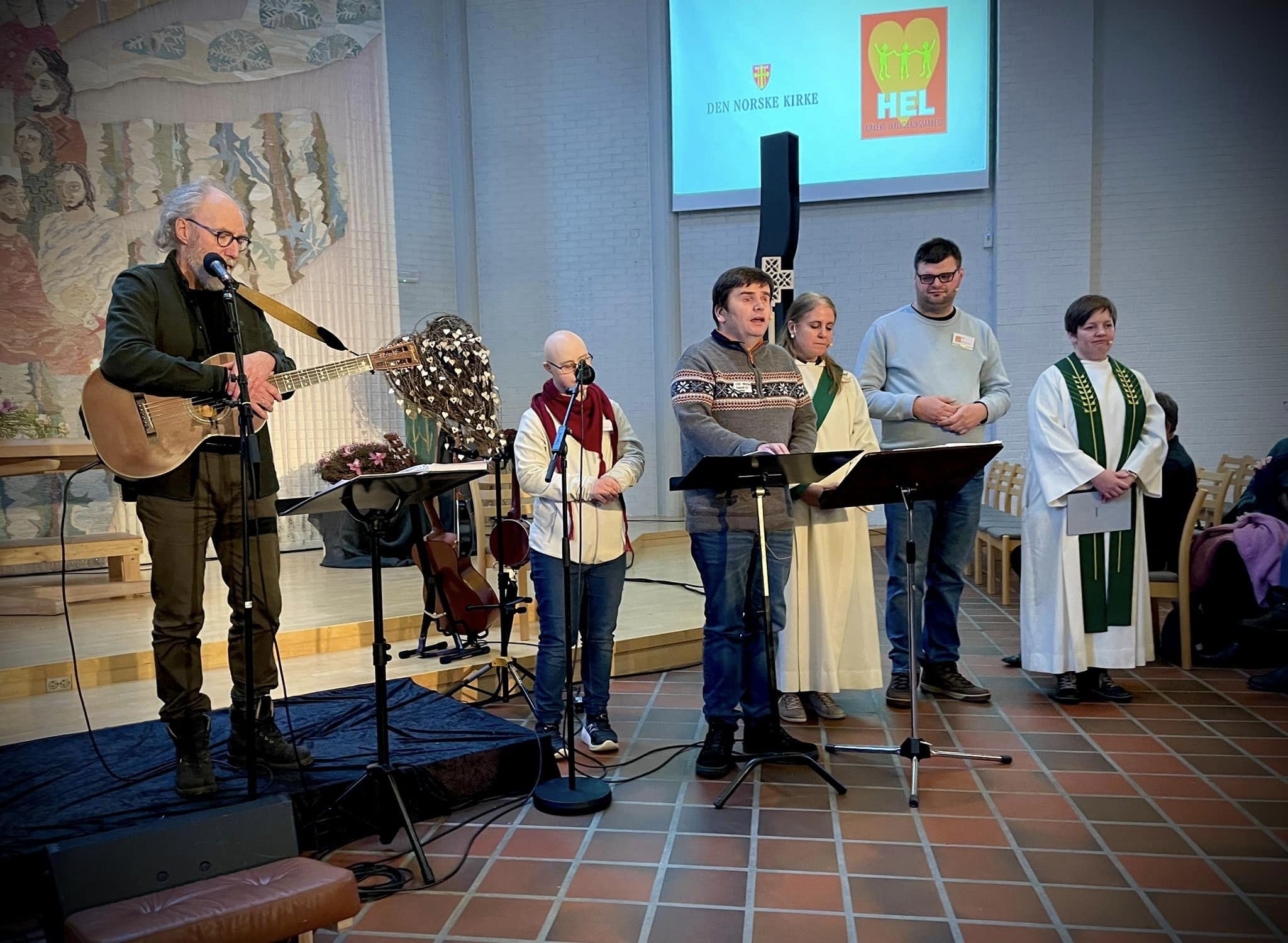 Seks personer står foran i en kirke. En mann har med seg gitar. To har prestekjoler