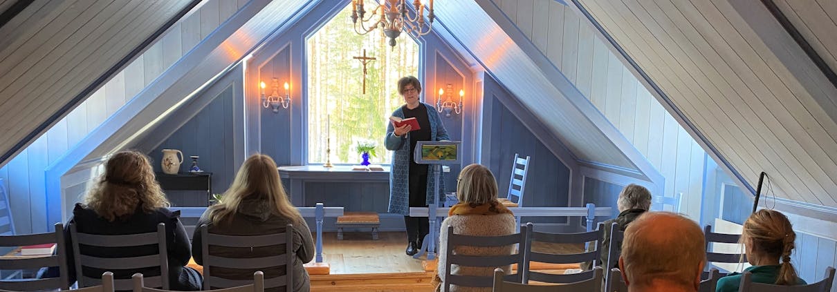 En person står foran i et kapell og leser en tekst. Det sitter 6 personer i benkeradene