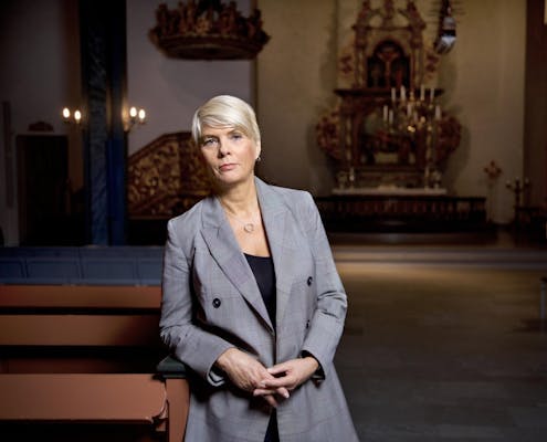 Kirkerådsleder Kristin Gunleiksrud Raaum står foran i en kirke, hun lener seg mot en kirkebenk og ser inn i kameraet