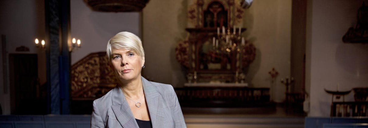 Kirkerådsleder Kristin Gunleiksrud Raaum står foran i en kirke, hun lener seg mot en kirkebenk og ser inn i kameraet