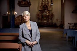 Kirkerådsleder Kristin Gunleiksrud Raaum står foran i en kirke, hun lener seg mot en kirkebenk og ser inn i kameraet