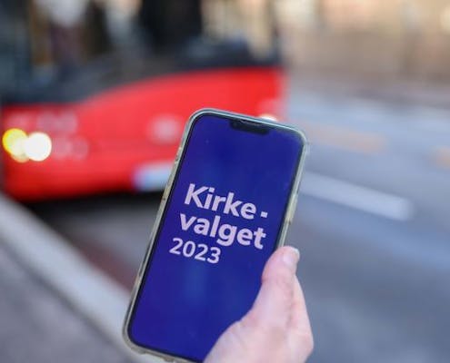 En mobiltelefon holdes av en person. På skjermen står det kirkevalget 2023