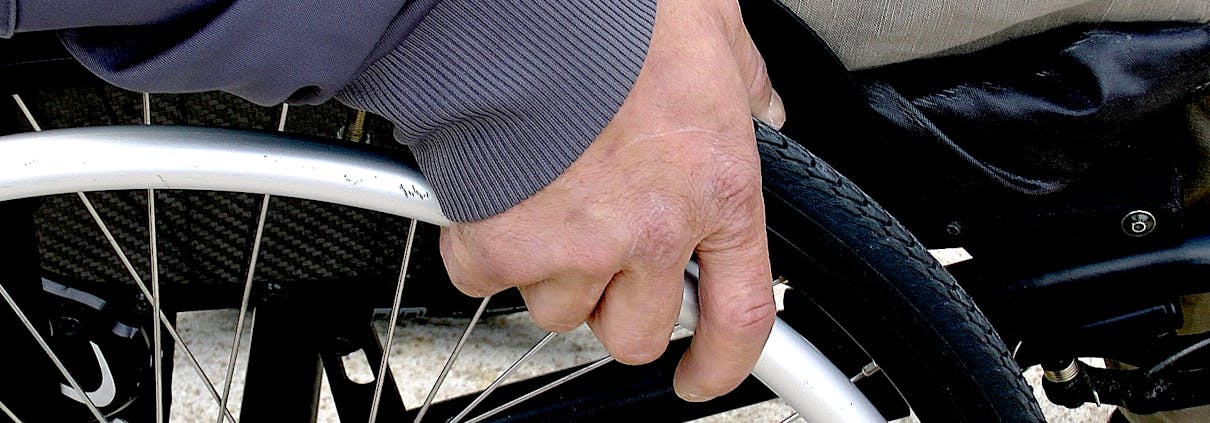 En person sitter i rullestol. Vi ser en hånd som holder på det ene hjulet.