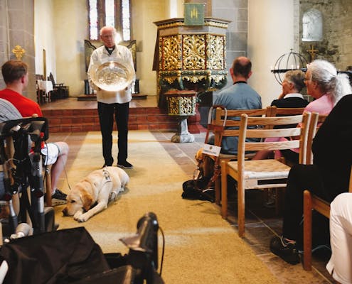 En gruppe mennesker sitter i benkeradene i en kirke. Foran står en prest og holder et metallfat
