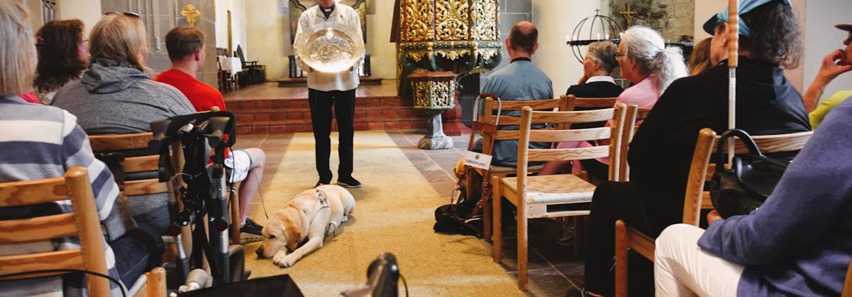 En gruppe mennesker sitter i benkeradene i en kirke. Foran står en prest og holder et metallfat