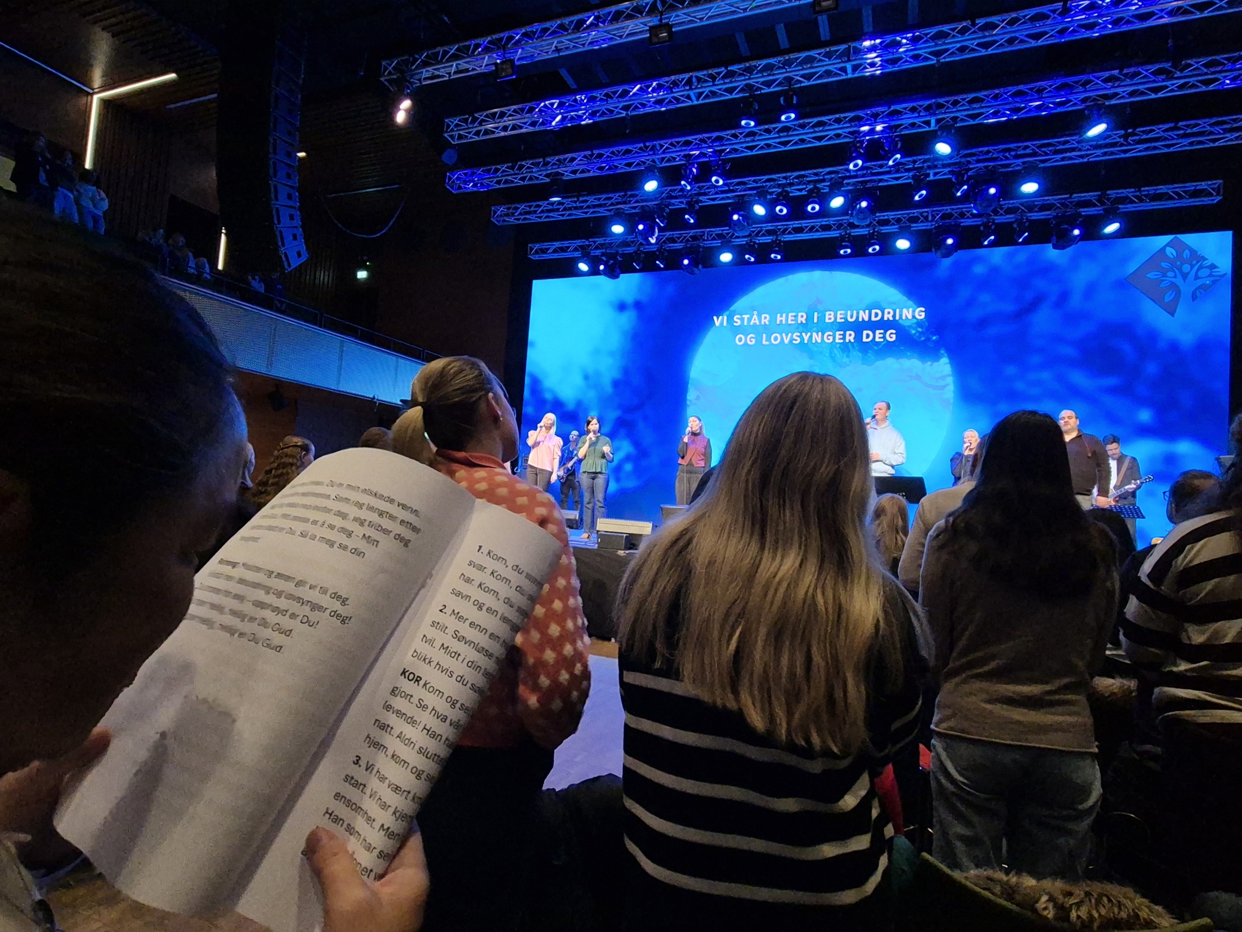 Bildet viser litt av et sanghefte i storskrift med forasmlingen foran og skrift på storskjerm bak forsangerne.